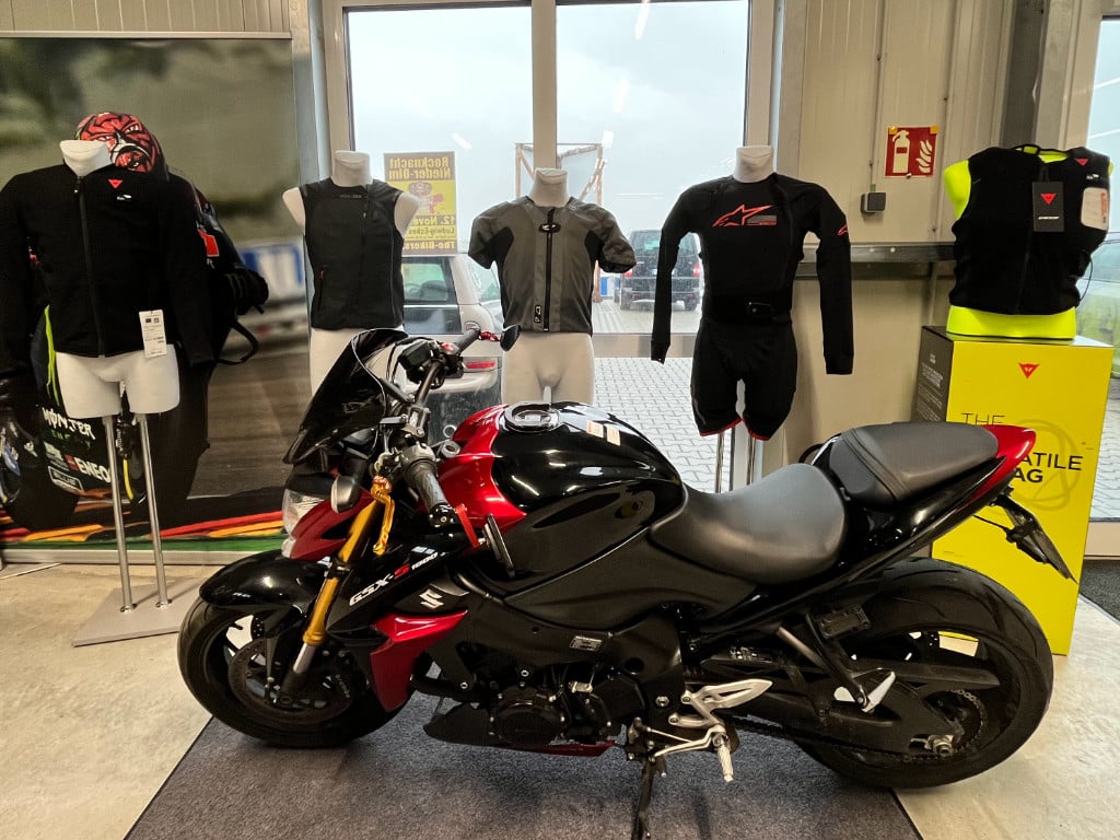 In einem Motorradzubehör-Shop sind verschiedene Airbag-Jacken und -Weste ausgestellt, während ein Motorrad im Vordergrund steht.