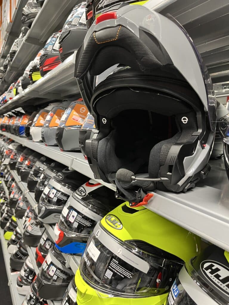 Ein grauer Motorradhelm ist auf einem Regal mit vielen weiteren Helmen platziert, die in verschiedenen Farben und Stilen angeordnet sind.